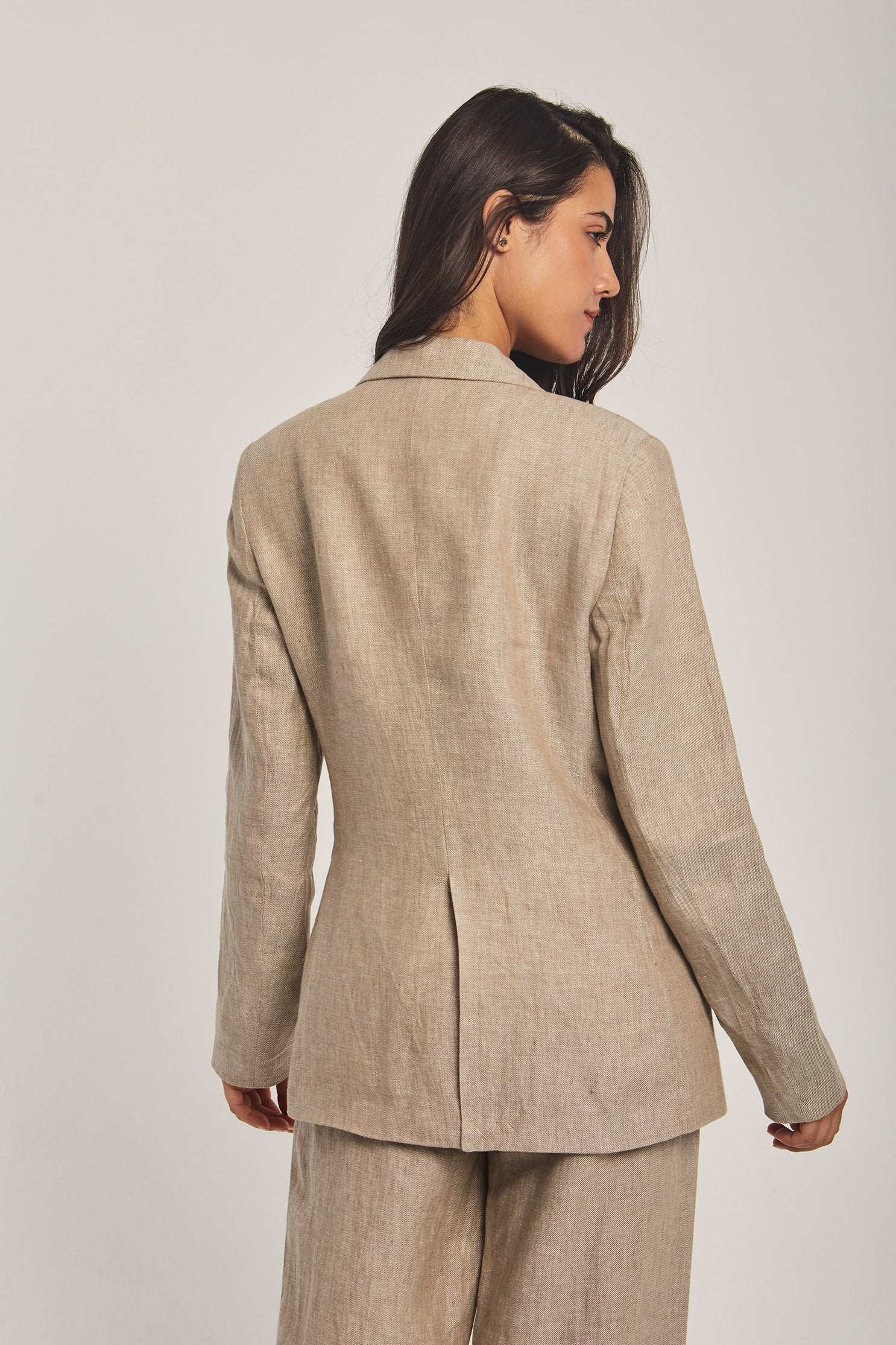 Woman wearing a beige linen blazer and trousers from Dalydress, back view against plain background