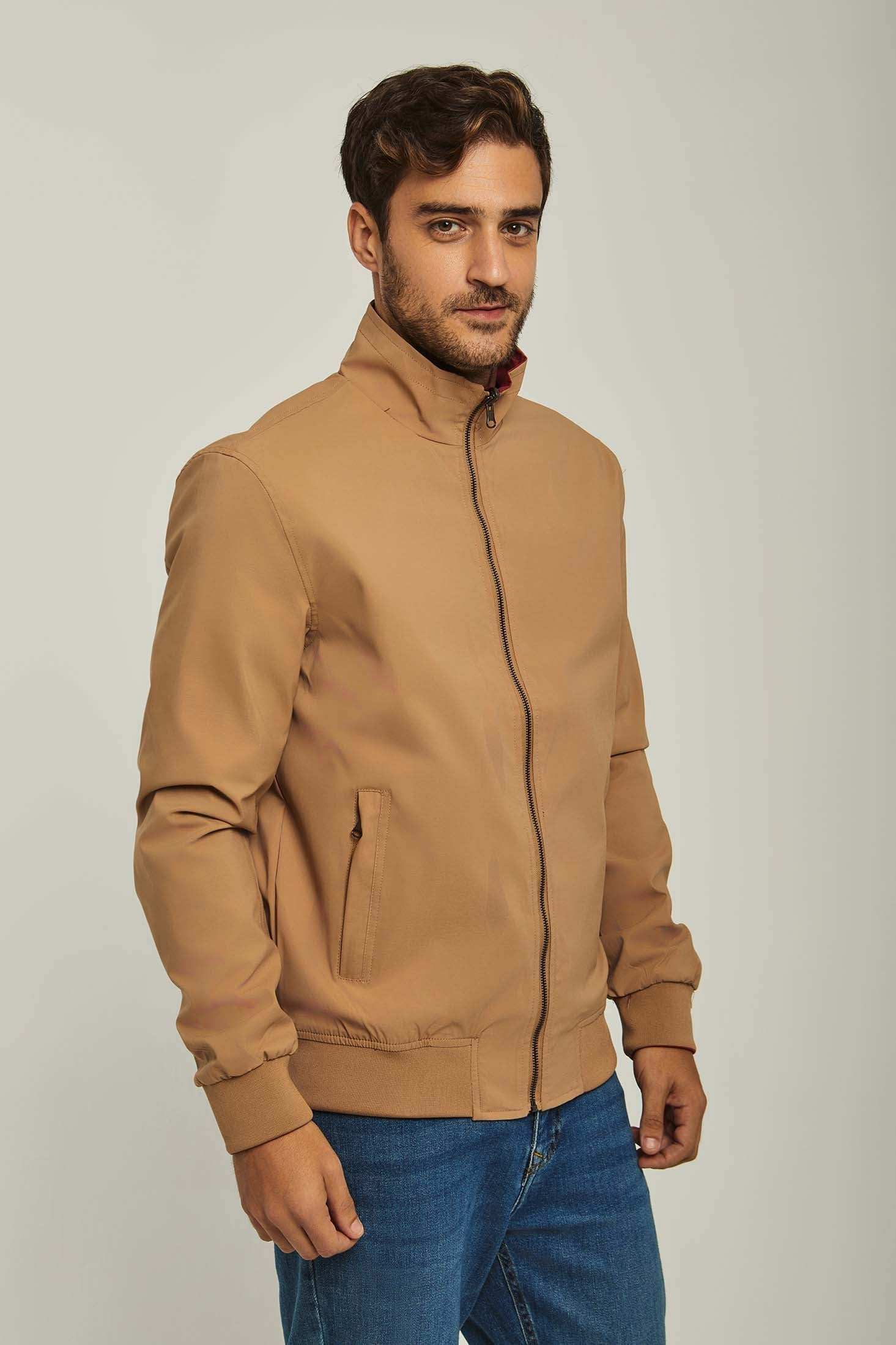 Man wearing tan zip-up jacket with stand collar and blue jeans on neutral background