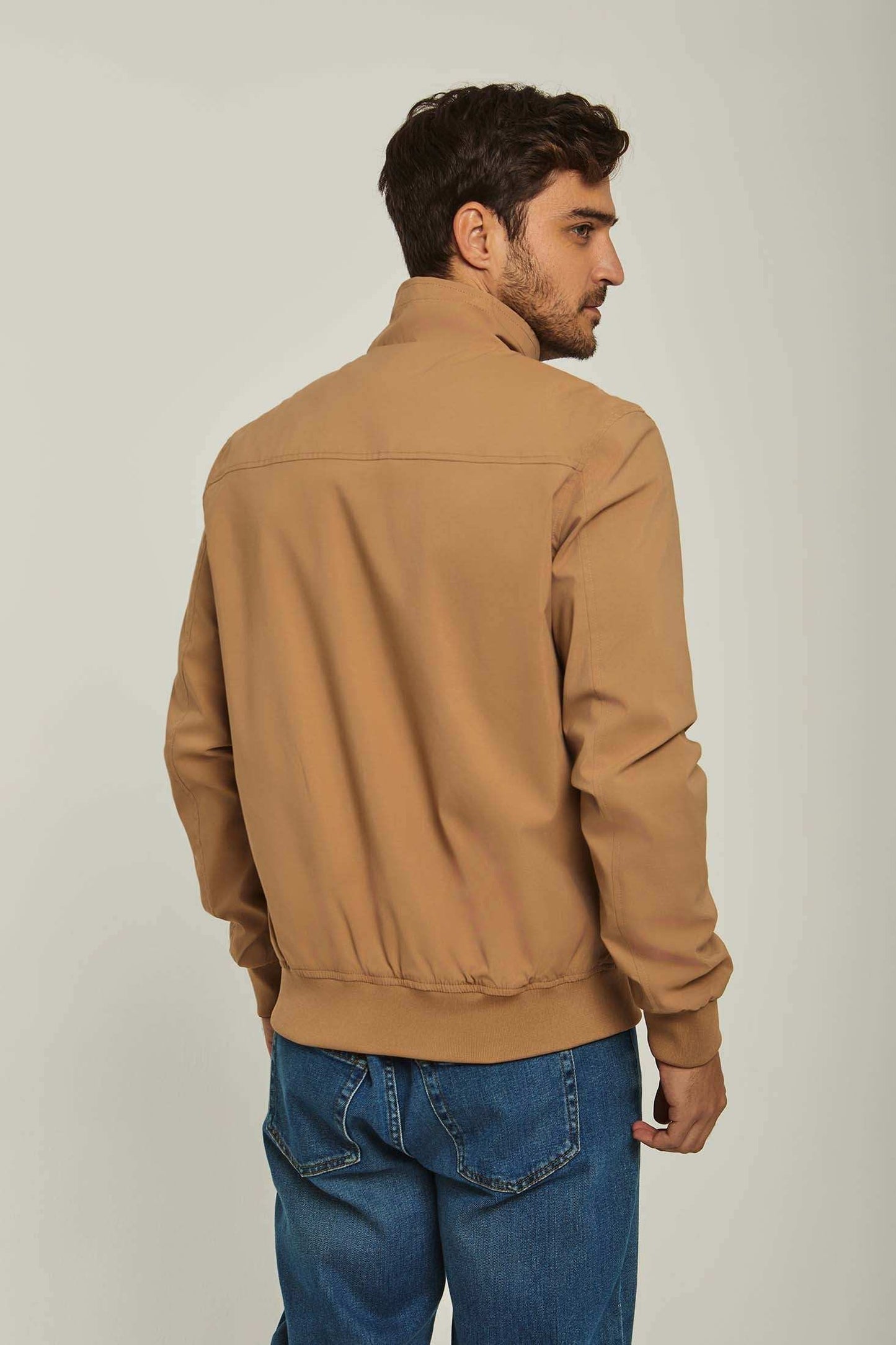 Man wearing a tan casual jacket and blue jeans, shown from the back on a neutral background.