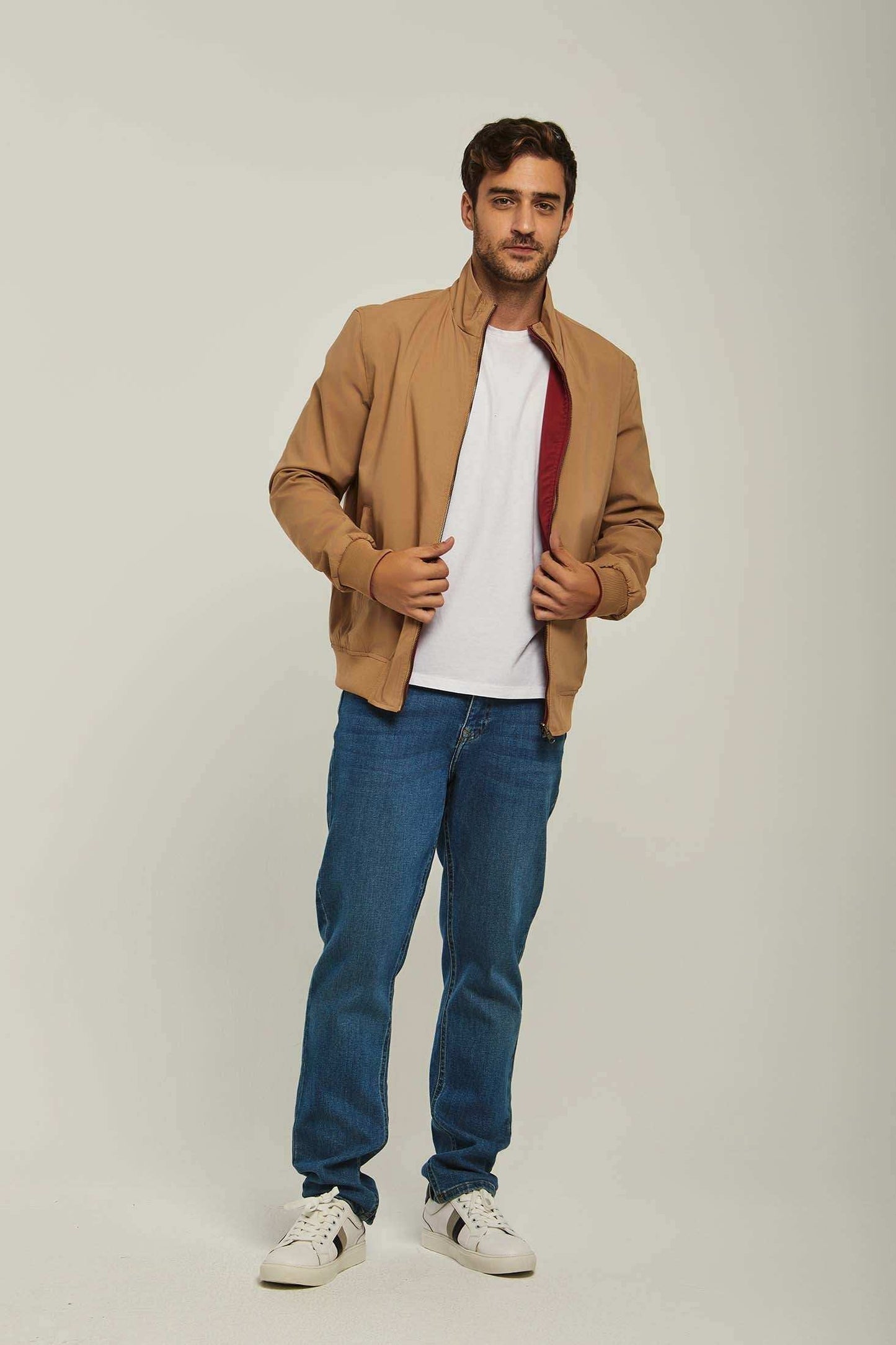 Man in casual tan jacket, white t-shirt, blue jeans, and white sneakers standing