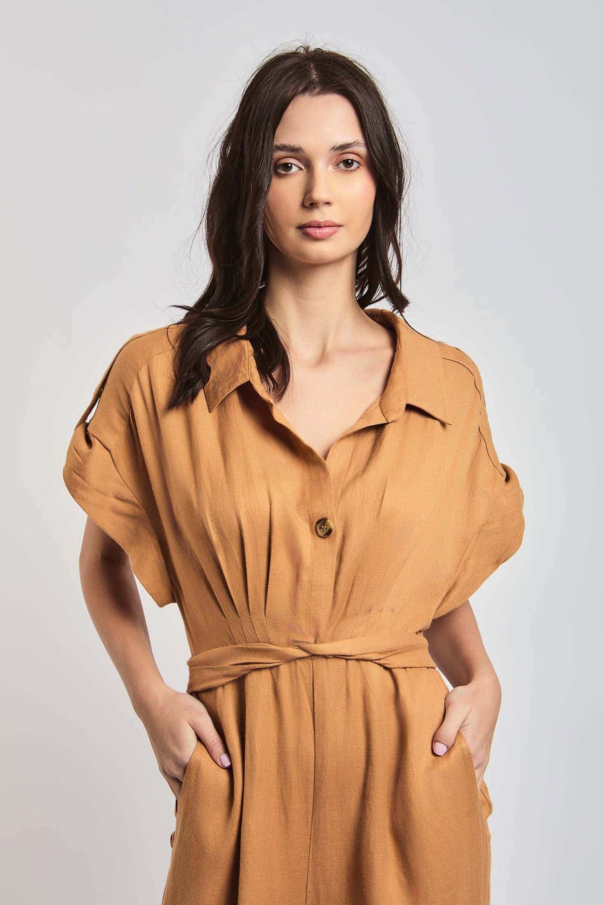 Woman in tan belted short sleeve shirt dress with collar, hands in pockets, neutral background