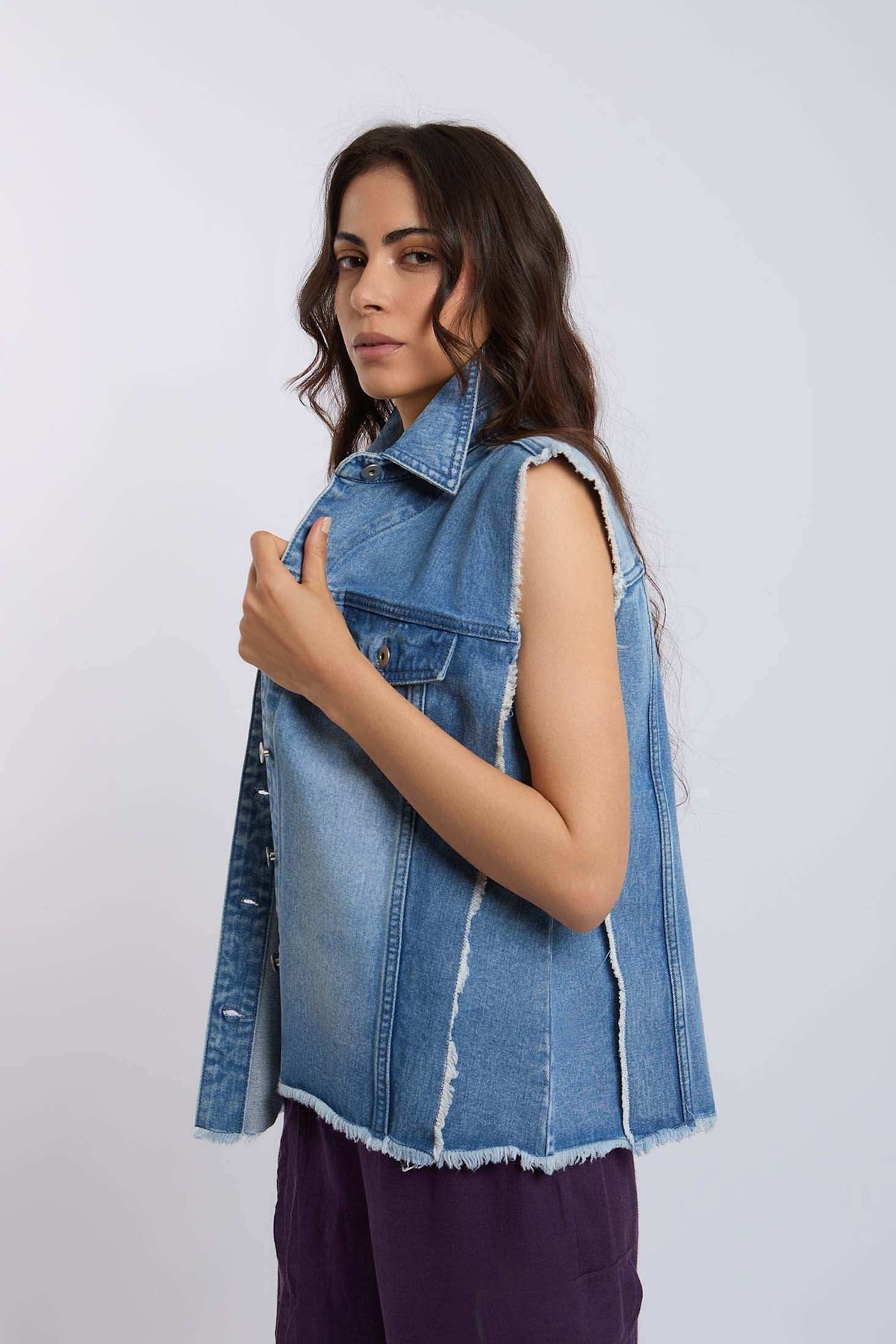Woman wearing sleeveless frayed blue denim vest and purple pants on white background