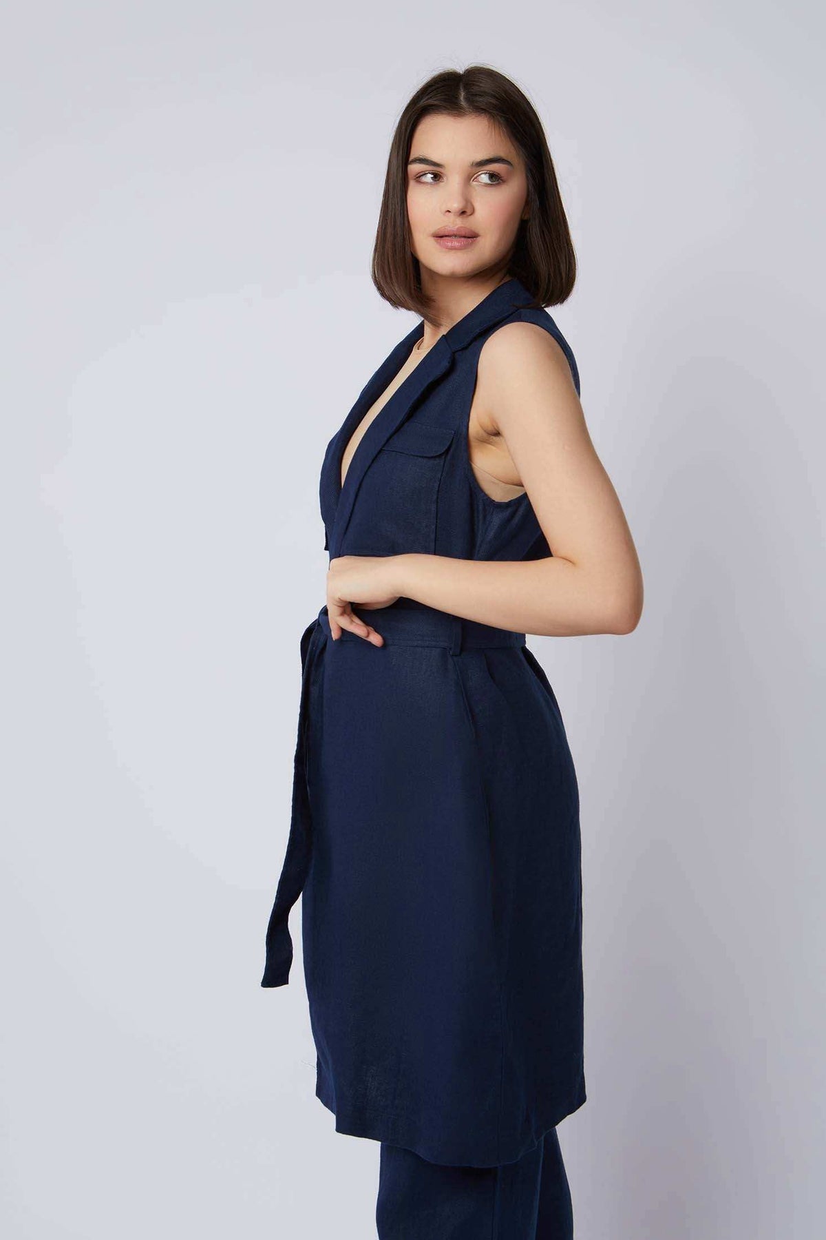 Woman wearing sleeveless navy blue belted vest with deep V-neck, posing sideways