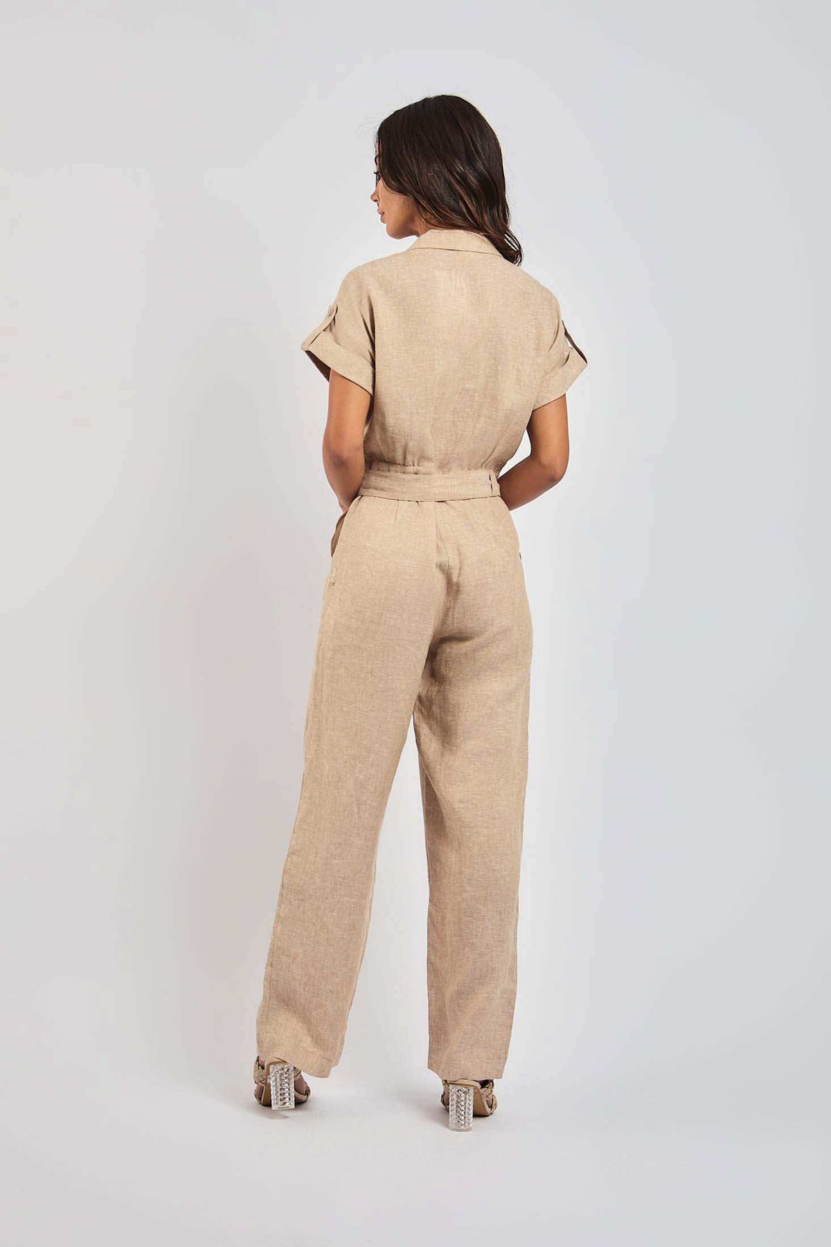 Woman wearing beige short sleeve belted linen jumpsuit, standing with back view