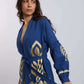 Woman wearing blue belted jacket with gold geometric embroidery, standing against light background
