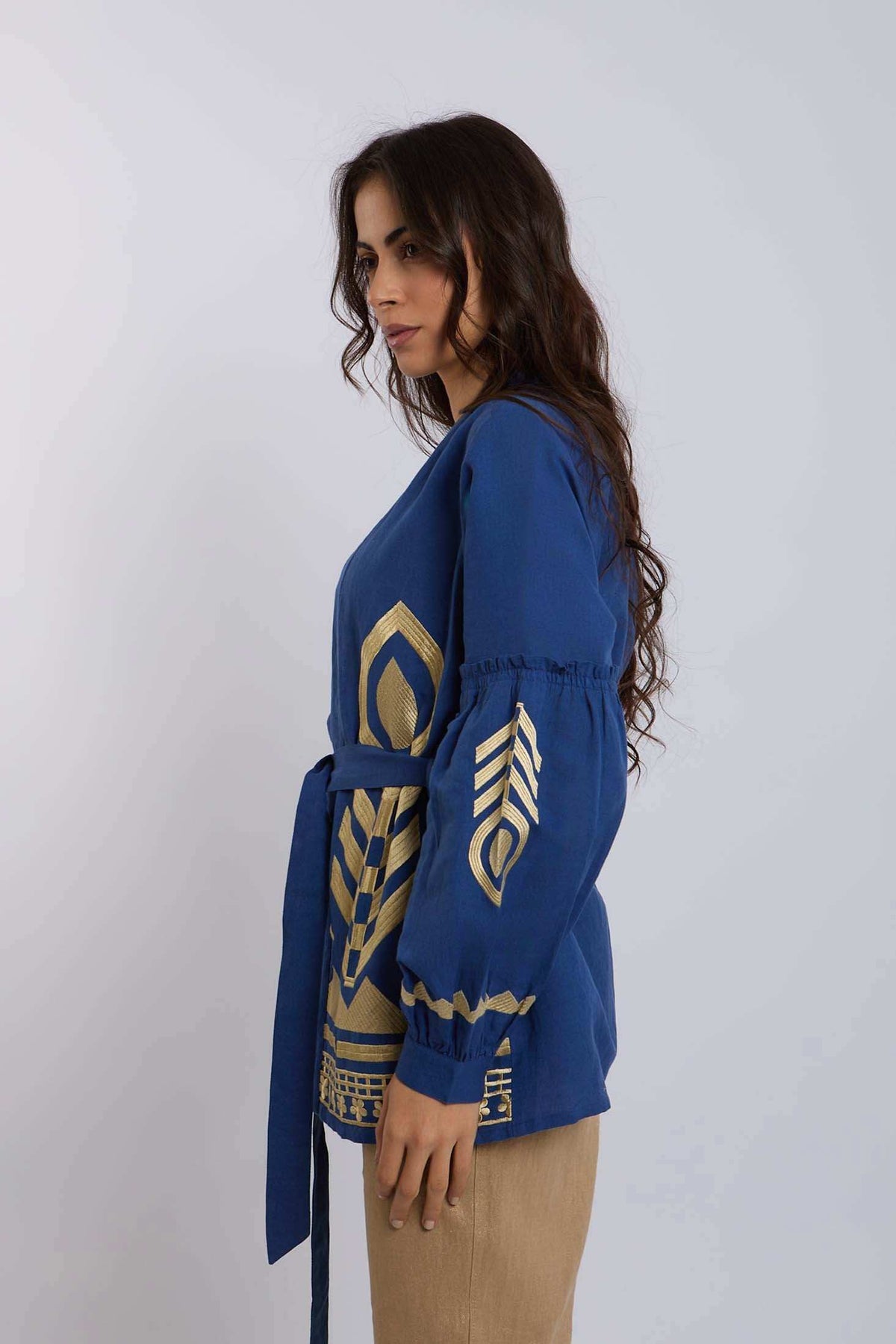 Woman in blue long-sleeve blouse with gold geometric patterns and waist belt, side view