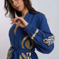 Woman wearing a blue belted dress with gold embroidered patterns on sleeves and waist