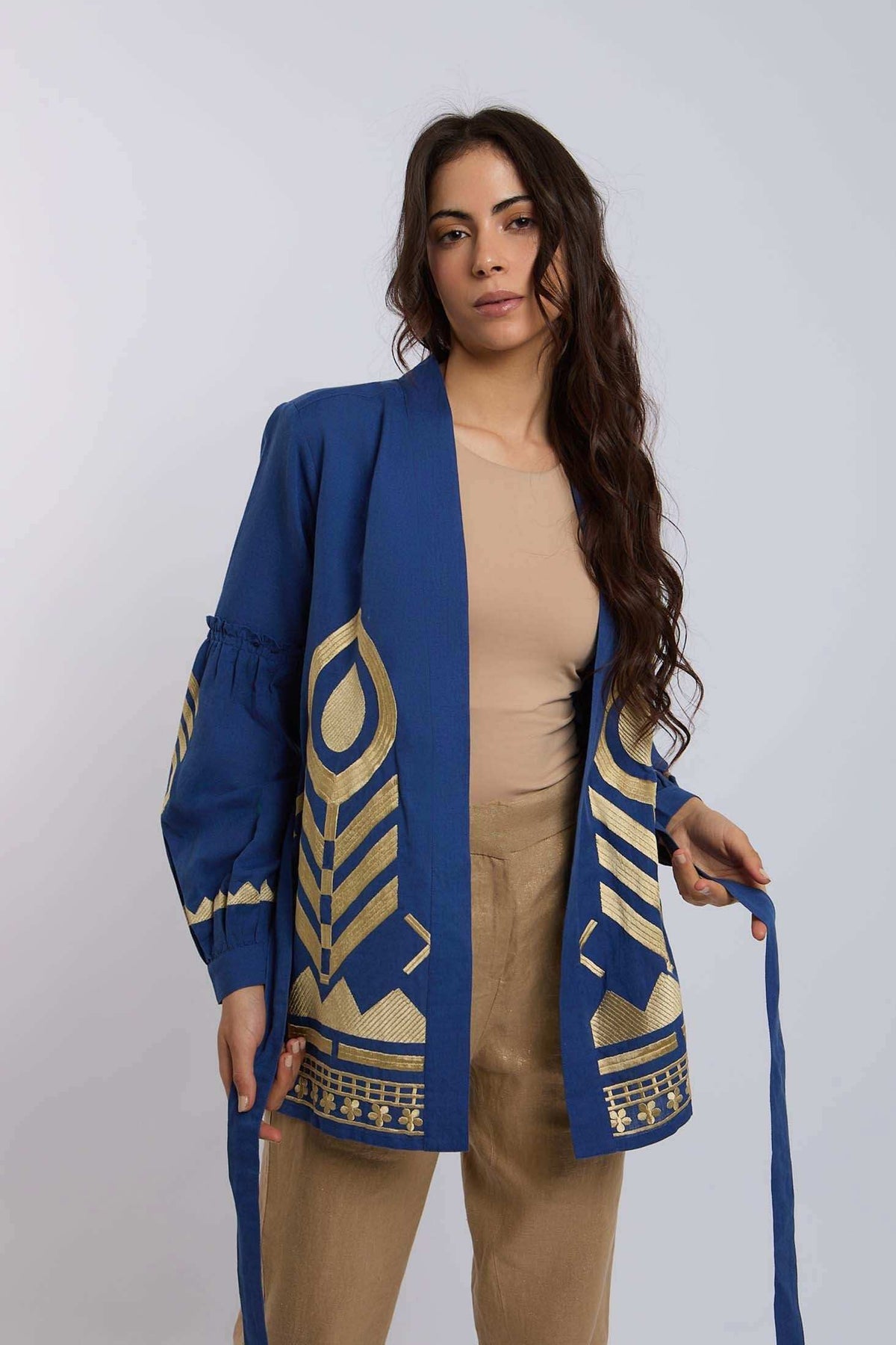 Woman wearing blue open-front jacket with gold geometric embroidery, beige shirt and pants