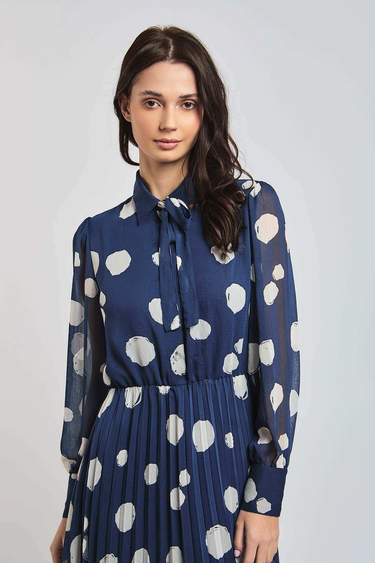 Woman wearing a Dalydress blue polka dot pleated dress with tie-neck detail