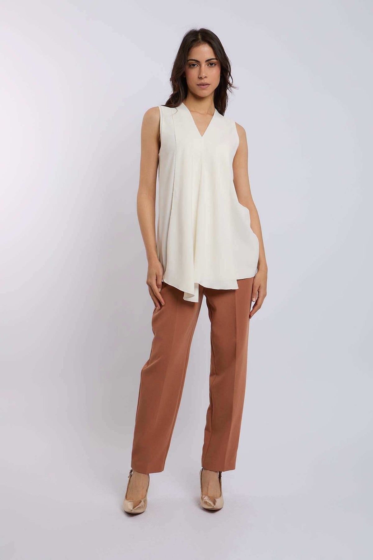 Woman wearing sleeveless cream tunic top and brown straight-leg trousers, standing indoors