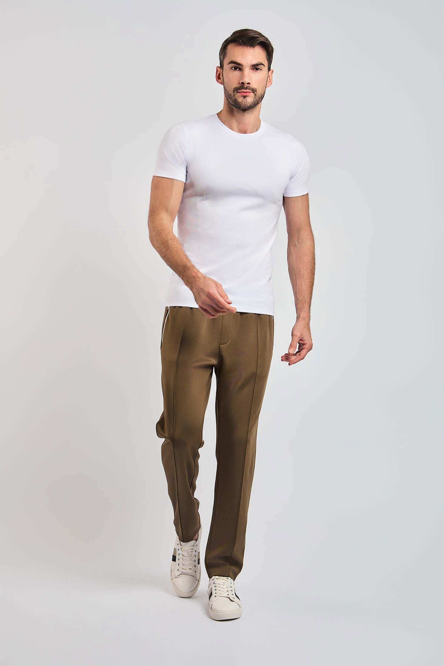Man in white t-shirt, brown tailored pants, and white sneakers on plain background
