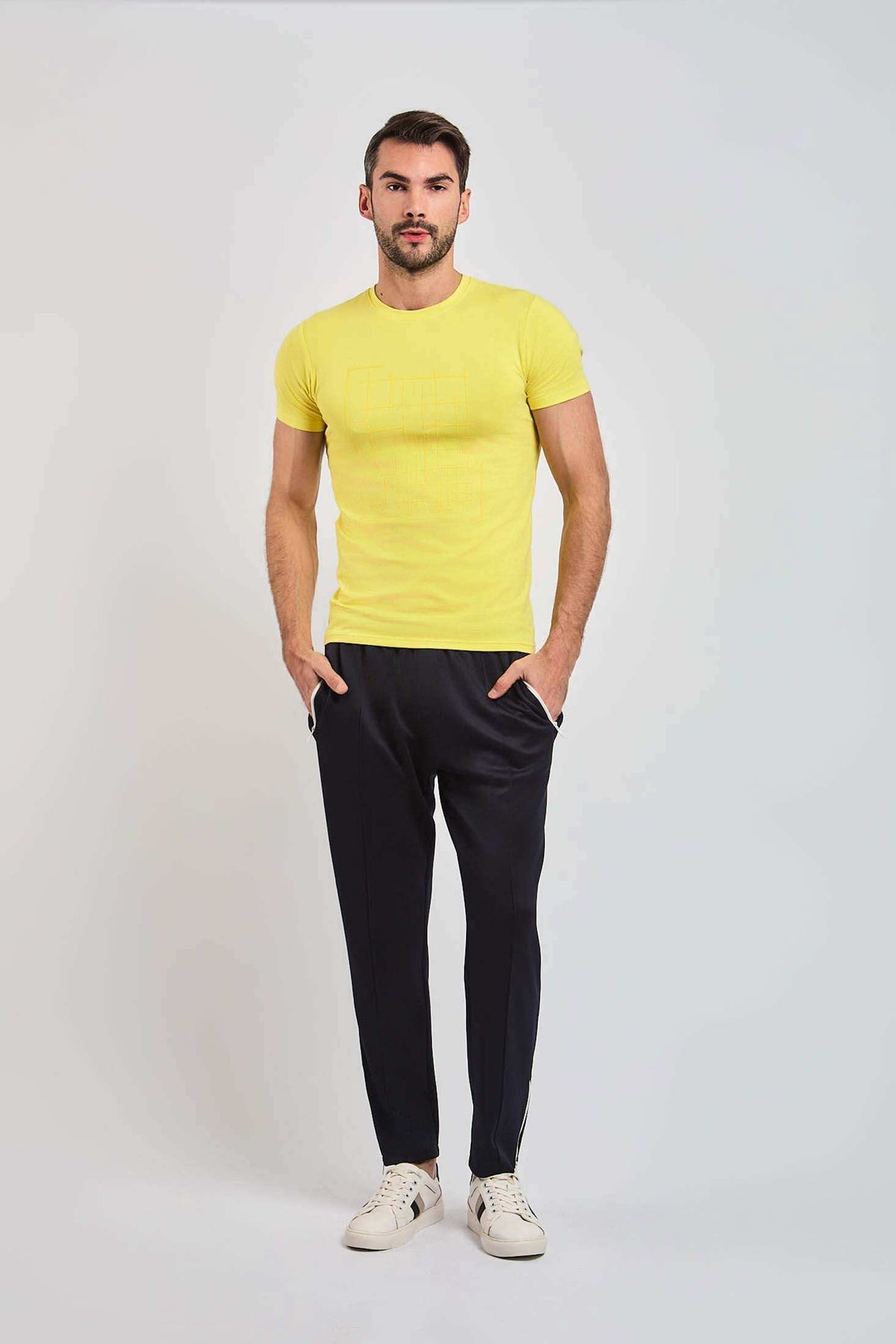 Man in a yellow t-shirt, black pants, and white sneakers posing on a plain background