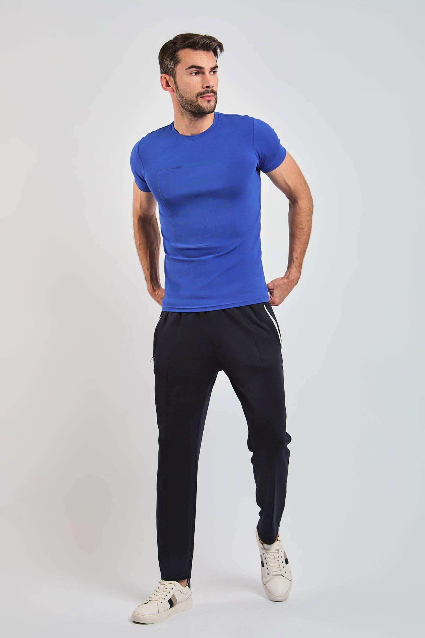 Man wearing a fitted blue t-shirt, black casual pants, and white sneakers, standing against plain background