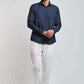 Man wearing a dark denim button-up shirt, white pants, and white sneakers against plain background