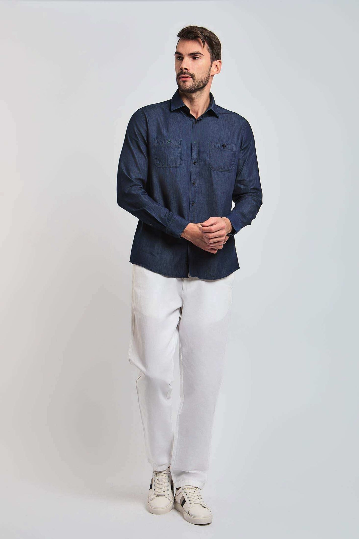 Man wearing a dark denim button-up shirt, white pants, and white sneakers against plain background
