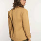Woman in tan blazer and green pants, posing from the back against white background