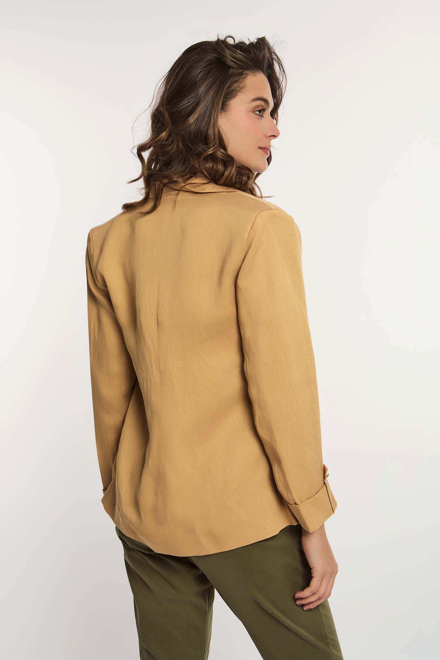 Woman in tan blazer and green pants, posing from the back against white background