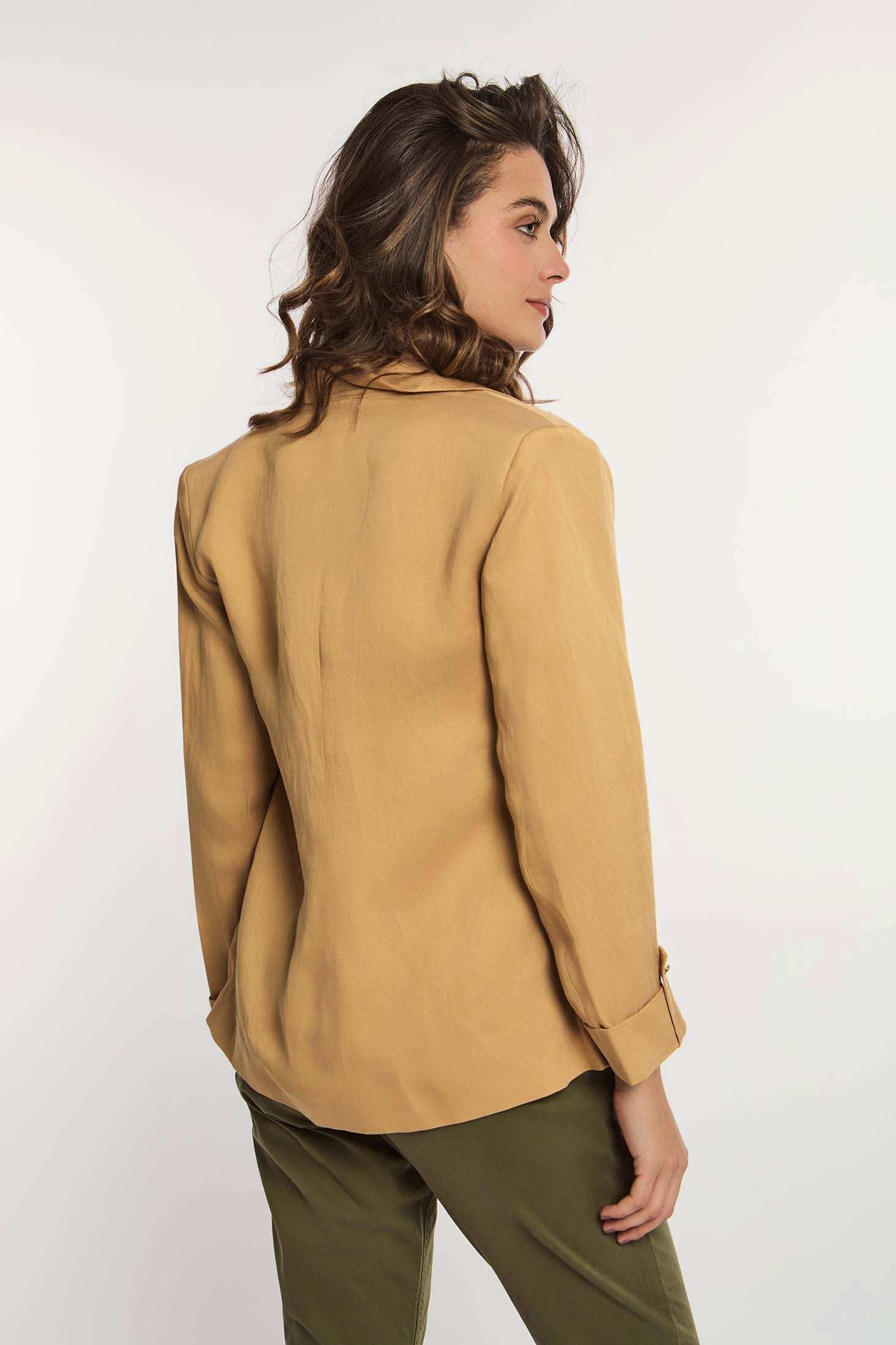 Woman in tan blazer and green pants, posing from the back against white background