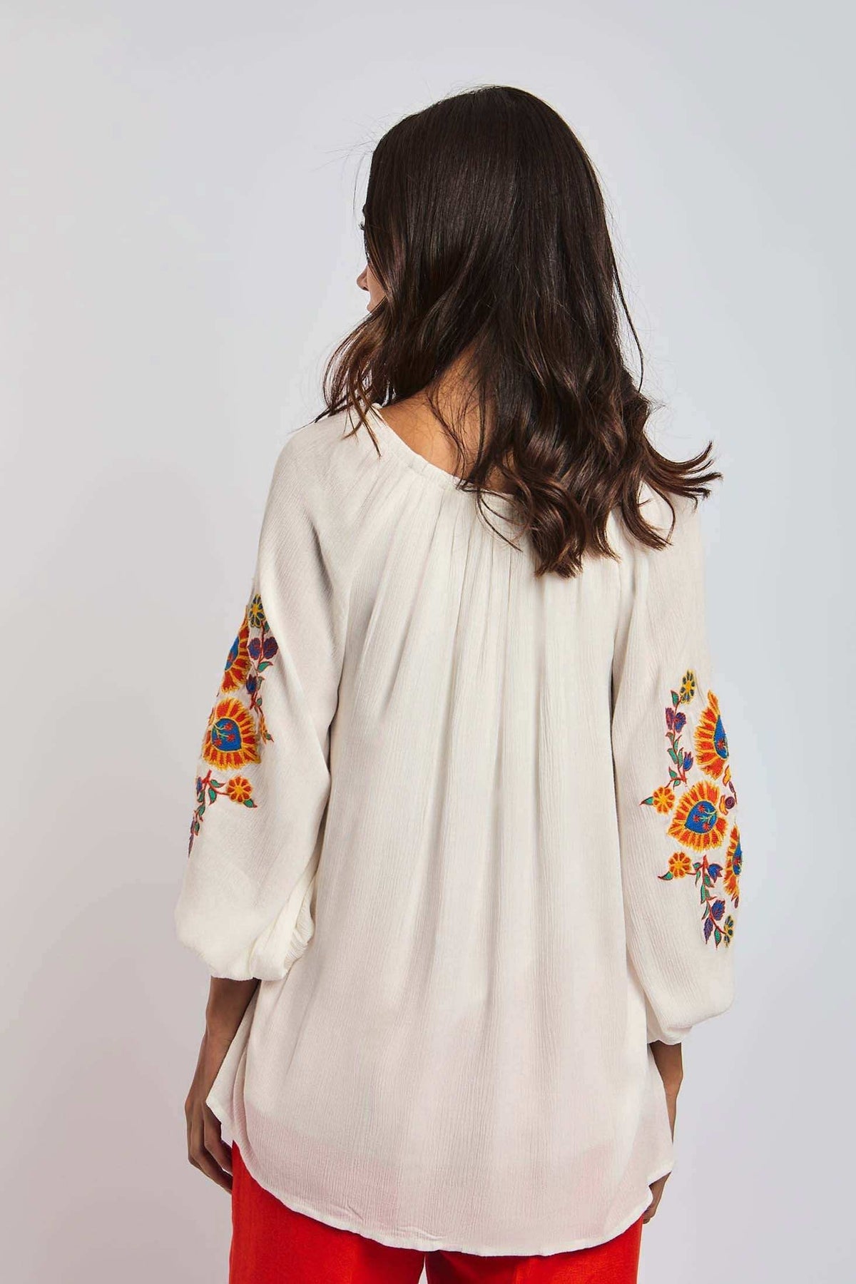 Woman wearing white blouse with colorful floral embroidery on sleeves, red pants, back view