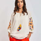 Woman wearing a white embroidered blouse with colorful tassels and bright orange pants