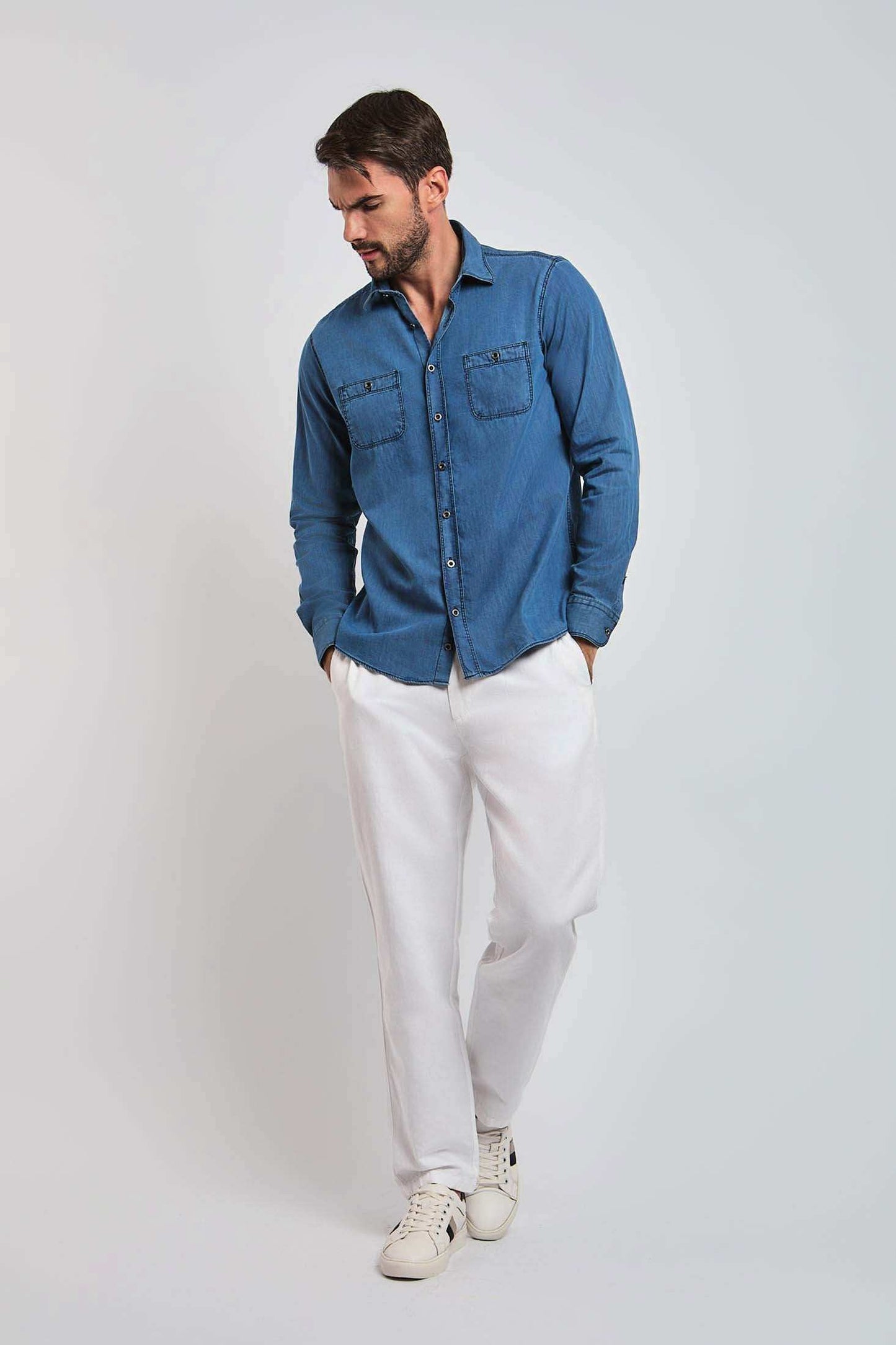 Man wearing blue denim shirt with chest pockets, white pants, and sneakers against plain background